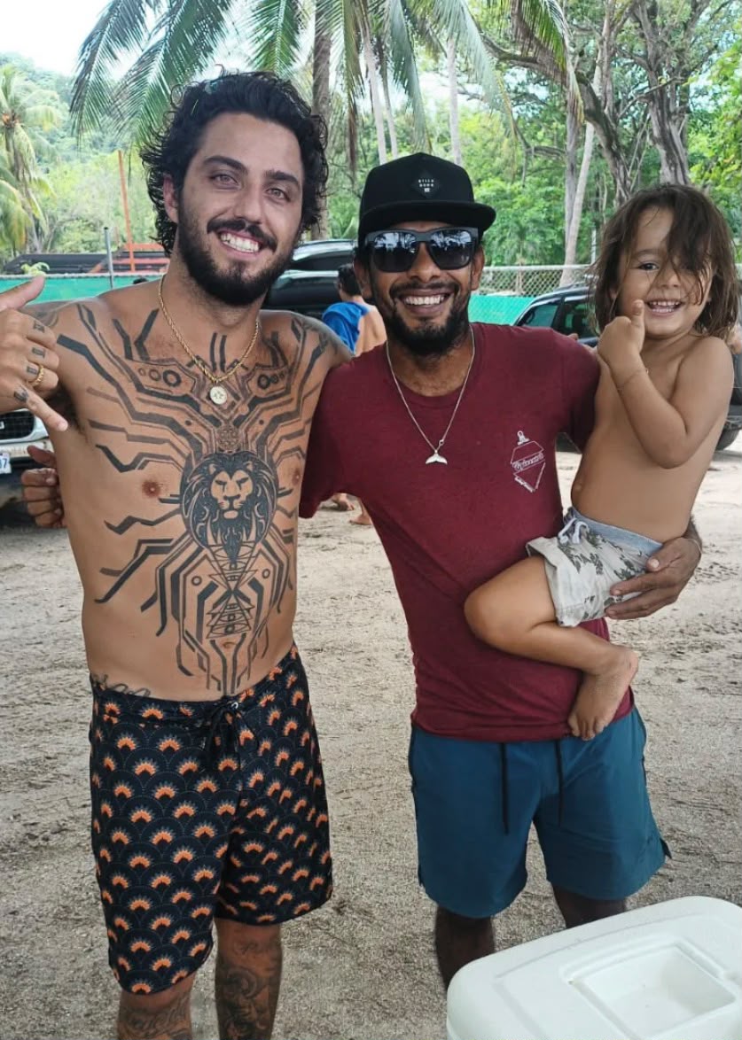 Papi holding his kid on the beach next to surfer Filipe Toledo, two-time world champion